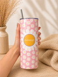 A hand holds a tall insulated tumbler with a pink floral print and a yellow circular label reading 'Ashleigh'. The tumbler has a clear lid and a metal straw, set on a wooden surface with dried flowers and a beige knit blanket in the background.