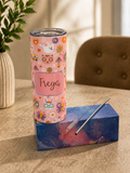 A pink insulated tumbler with a colorful butterfly and floral pattern and the name 'Freyra' printed on the front, displayed on top of a blue galaxy-patterned box with a metal straw, set on a tabletop in a softly lit indoor setting.