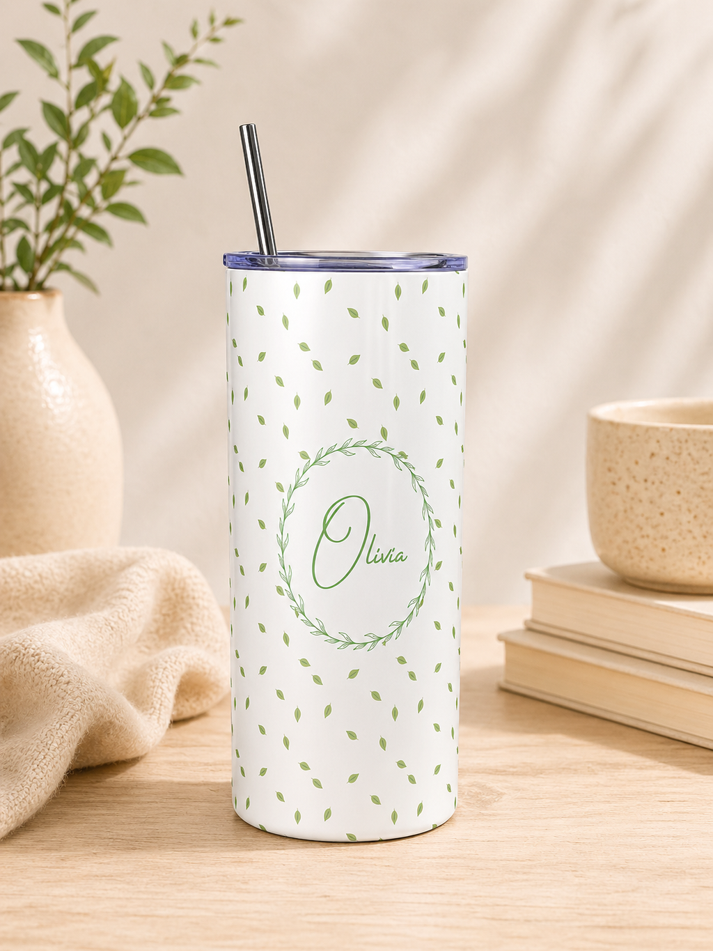 A tall insulated tumbler with a clear lid and metal straw, standing on a wooden surface. The tumbler is white with a scattered small green leaf pattern and a central green wreath outline containing the name 'Olivia' in script.