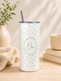 A tall insulated tumbler with a clear lid and metal straw, standing on a wooden surface. The tumbler is white with a scattered small green leaf pattern and a central green wreath outline containing the name 'Olivia' in script.
