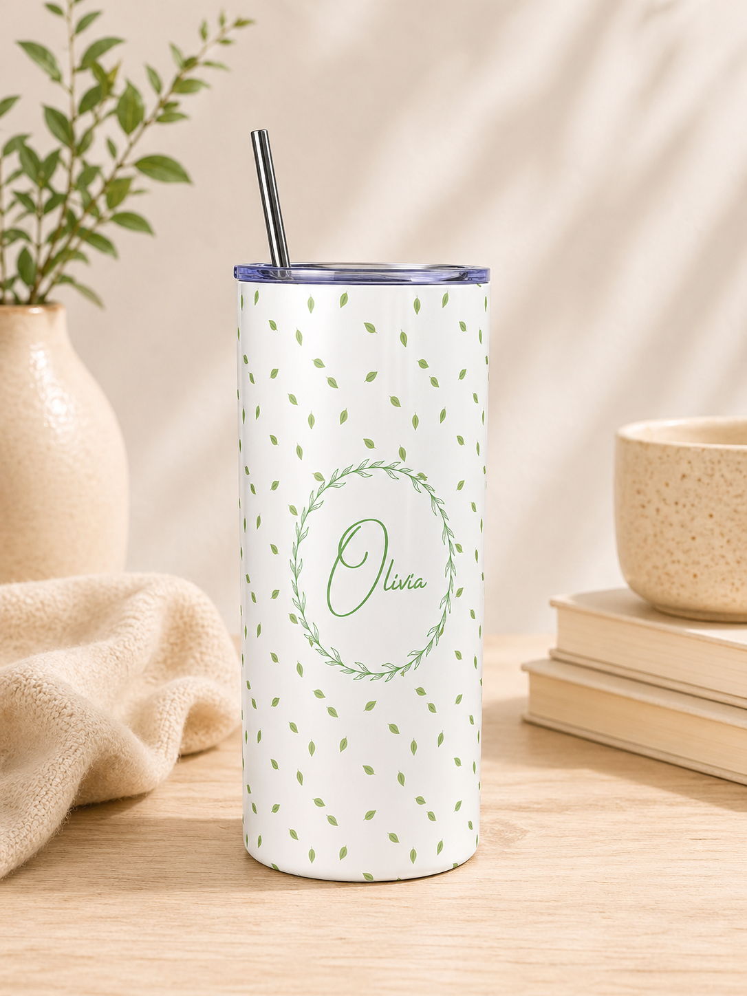 A tall insulated tumbler with a clear lid and metal straw, standing on a wooden surface. The tumbler is white with a scattered small green leaf pattern and a central green wreath outline containing the name 'Olivia' in script.