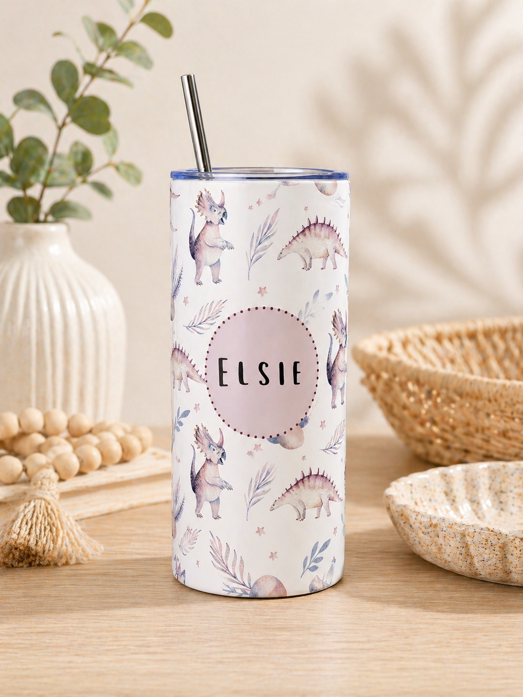 A tall insulated tumbler with a clear lid and metal straw, decorated with pastel dinosaur illustrations and the name 'Elsie' in the center, set on a wooden table with decorative props in a warm indoor setting.