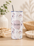 A tall insulated tumbler with a clear lid and metal straw, decorated with pastel dinosaur illustrations and the name 'Elsie' in the center, set on a wooden table with decorative props in a warm indoor setting.